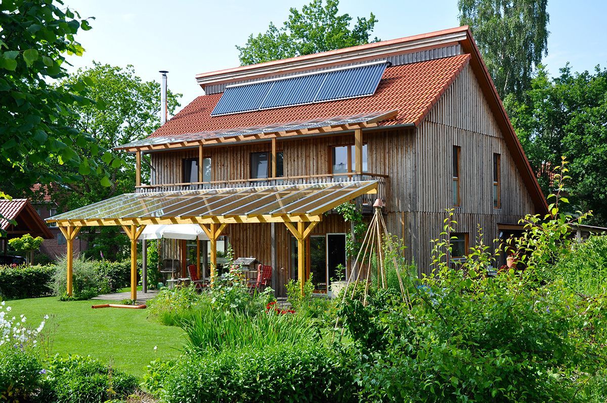 Modernes Holzhaus mit vertikaler Holzfassade, Solaranlage auf dem roten Ziegeldach und einer großen Glasüberdachung über der Terrasse in einem grünen Garten